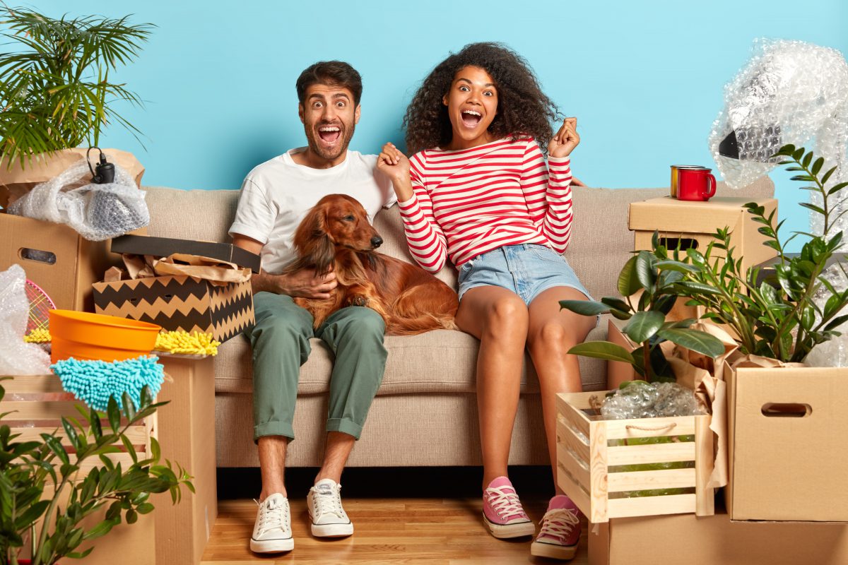 Extremely happy couple sat on a sofa with their dog surrounded by boxes